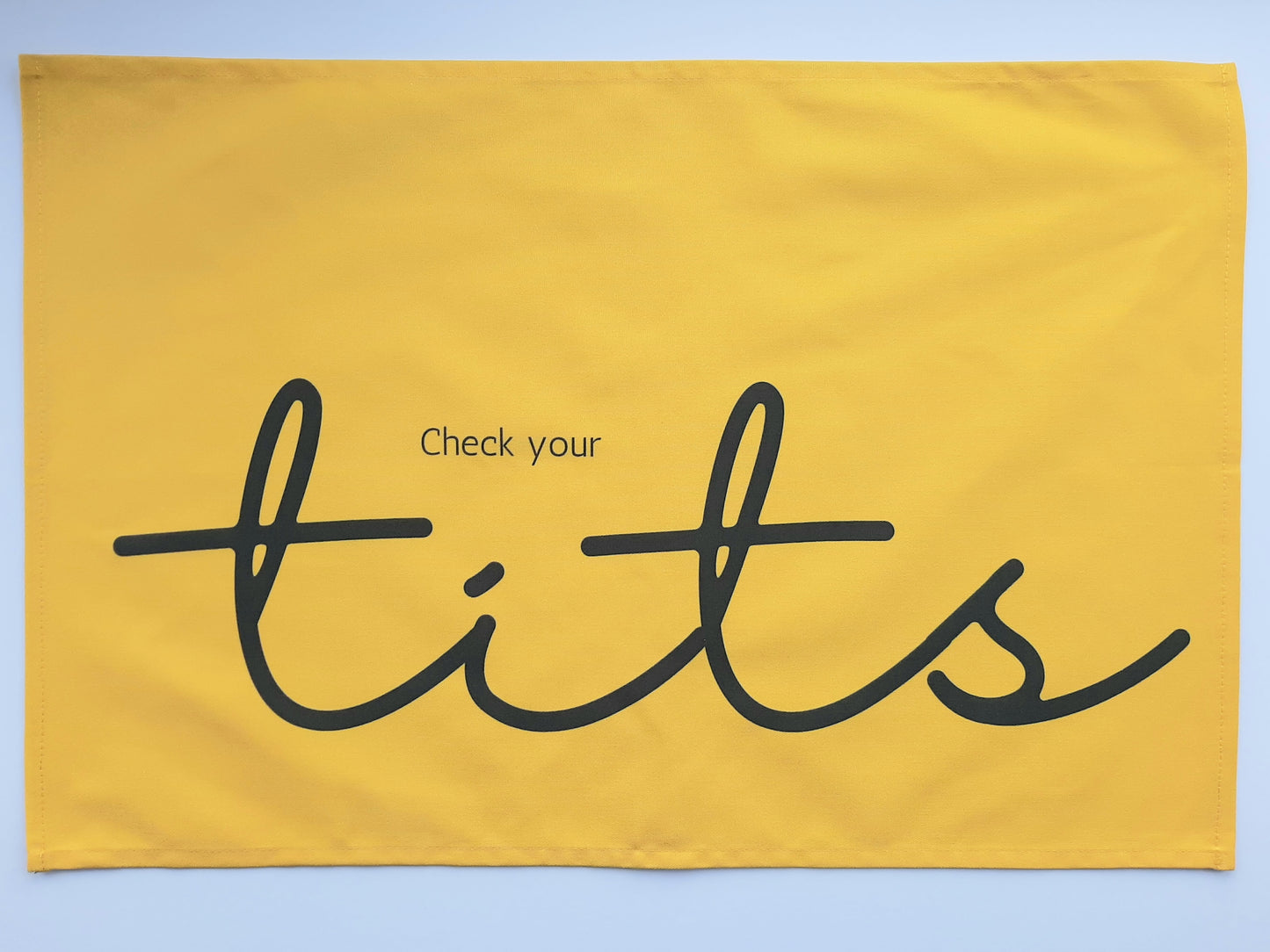 Photo of yellow tea towel with grey text reading Check your tits